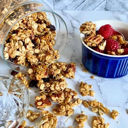 homemade granola in a glass container on its side dumped over with cup of raspberries